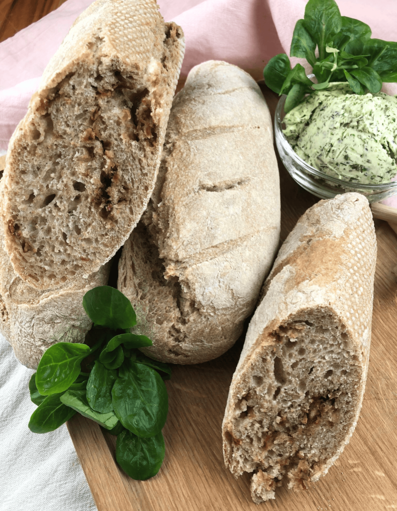 Nahaufnahme eines Zwiebelbaguettes: Die goldbraune Kruste zeigt leichte Risse, und die Röstzwiebeln verleihen dem Brot eine appetitliche Textur. Im Hintergrund ist eine Schale mit cremiger Kräuterbutter zu sehen.