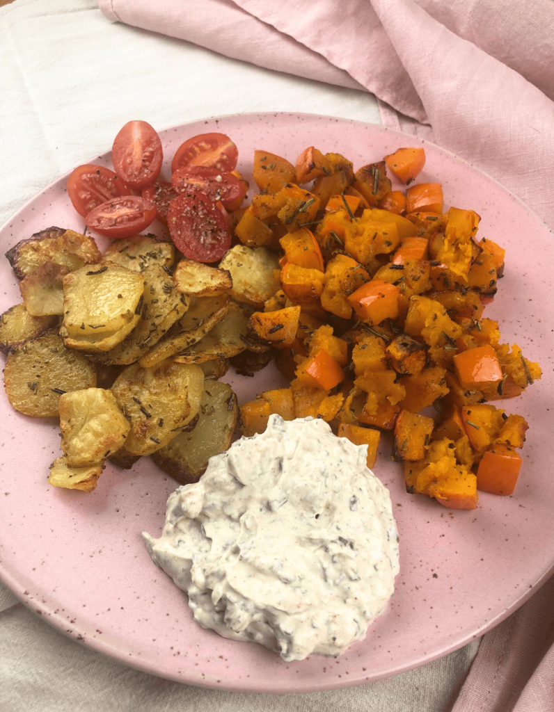 Ein buntes Gericht mit geröstetem Hokkaido-Kürbis und goldbraunen Kartoffelscheiben, serviert mit einem cremigen Kräuter-Dip mit halbierten Tomaten als Beilage.