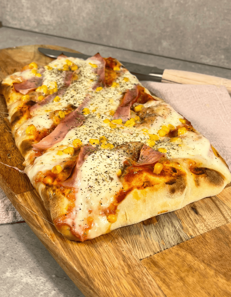 Frisch gebackene Pinsa: Rechteckiger Teigfladen, belegt mit zartem Kochschinken, geschmolzenem Mozzarella und goldbraunen Rändern.