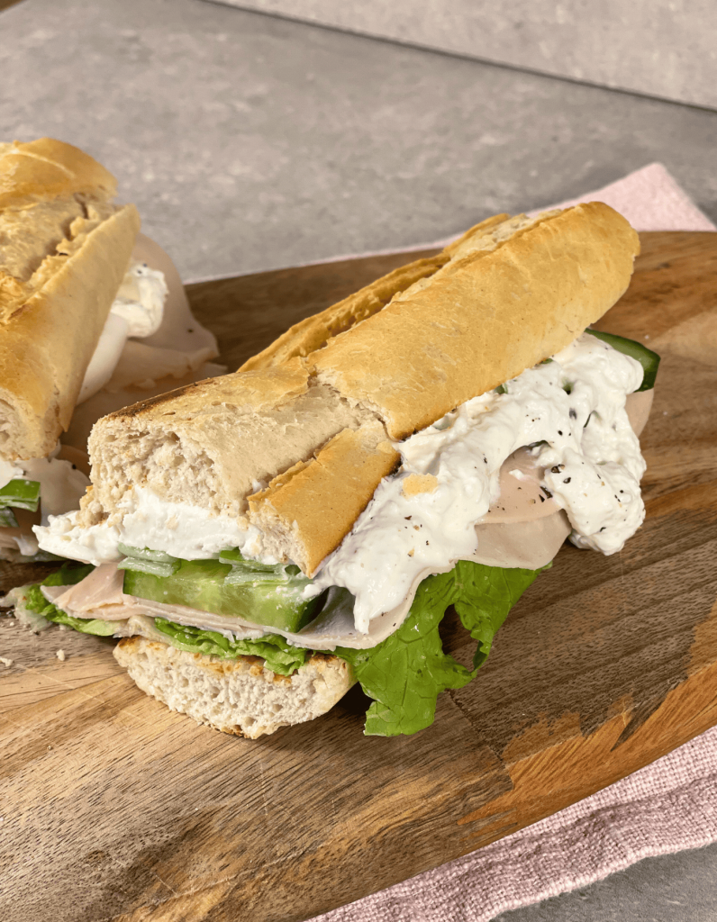 Nahaufnahme eines saftigen Baguettes, dessen Burrata zart schmilzt und die frischen Kräuter auf dem Holzbrett das Gesamtbild abrunden.