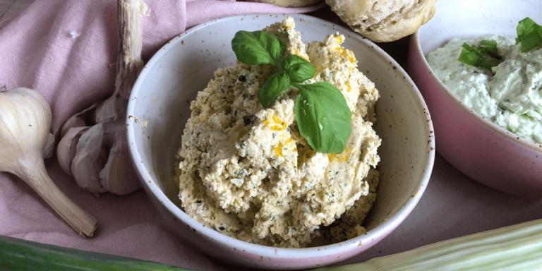 Der cremige Frischkäse-Dip, mit Basilikumblättern garniert, wird in einer eleganten Schale präsentiert. Dahinter liegt eine Knoblauchknolle, die den Fokus auf die natürlichen Zutaten lenkt.