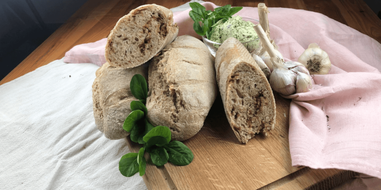 Frisch gebackenes Zwiebelbaguette liegt auf einem rustikalen Holzbrett. Die knusprige, goldbraune Kruste ist leicht eingerissen, und die Röstzwiebeln sind deutlich sichtbar. Daneben steht eine Schale mit selbstgemachter Kräuterbutter, dekoriert mit frischen Kräutern.
