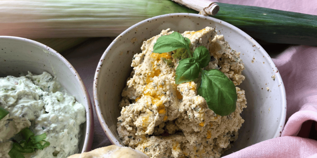 Eine Schale voller cremigem Dip, verziert mit frischem Basilikum. Im Hintergrund erkennt man eine Knoblauchknolle, die auf die Zutaten des Rezepts hinweist.
