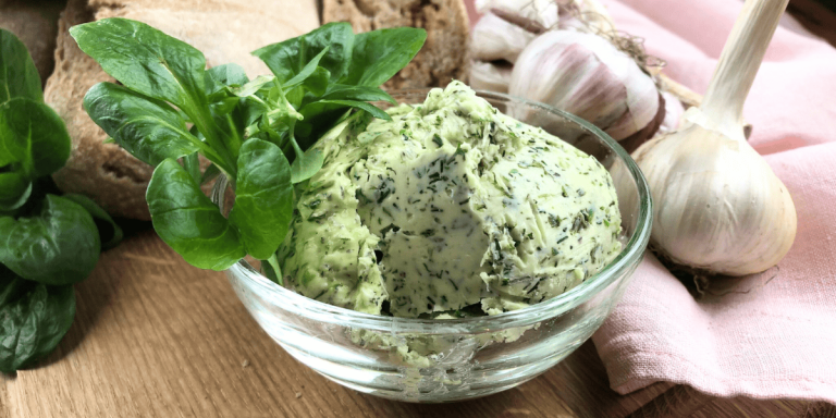 Eine Portion selbstgemachter Kräuterbutter auf einem kleinen Holzbrett. Die Butter zeigt deutlich die grünen Kräuter und Knoblauchstückchen, dekoriert mit frischer Kräutern.