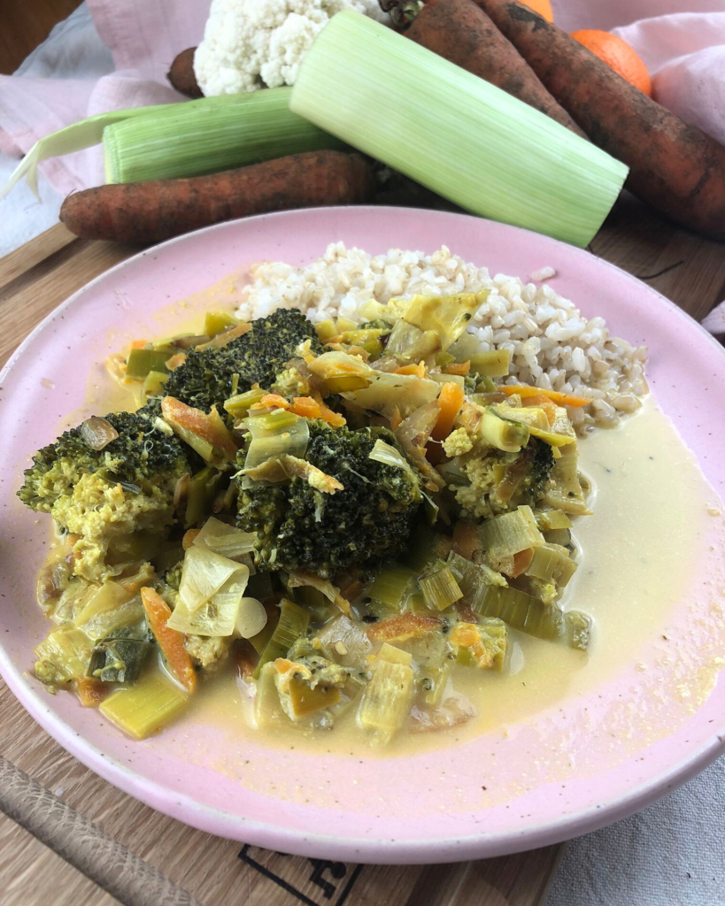 Bunter Genuss: Cremiges Curry-Gemüse angerichtet auf einem rosa Teller mit frischem Porree und Möhren-Deko. 