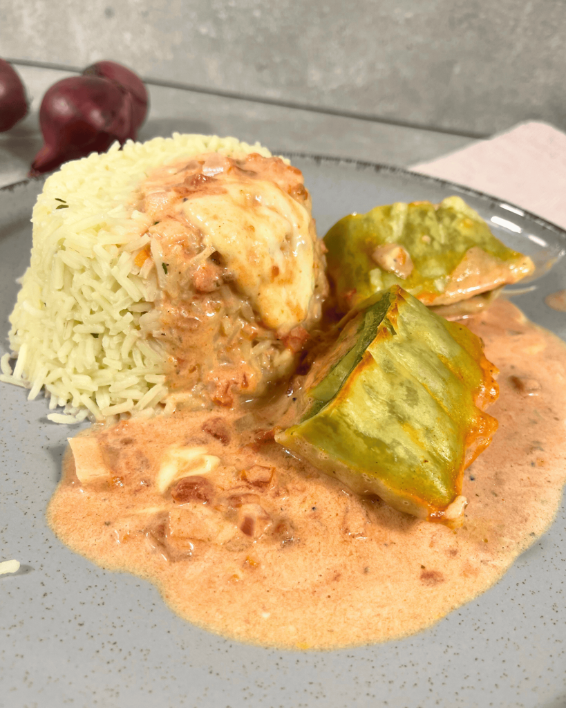 Ein Teller mit hellem Reis, cremiger hellroter Tomatensoße und saftigen Maultaschen, dekorativ angerichtet. Im Hintergrund liegen rote Zwiebeln, die das Gesamtbild abrunden.