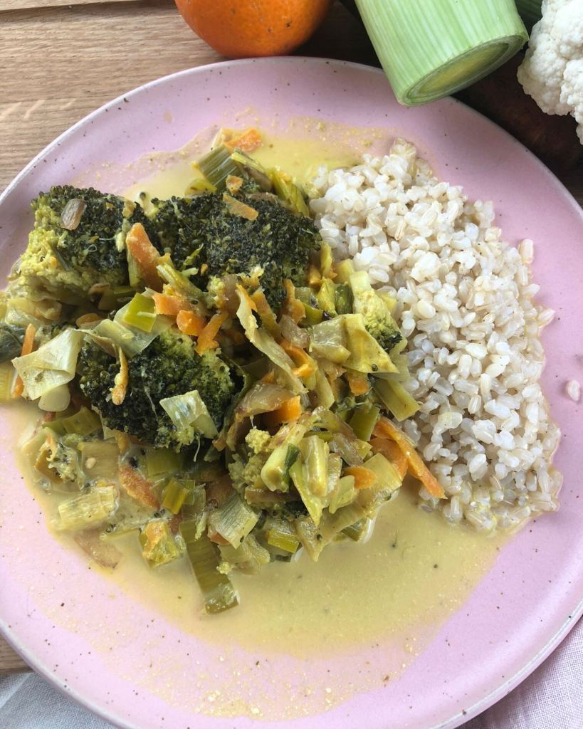 Fruchtiges Curry-Gemüse serviert auf einem rosa Teller – im Hintergrund frischer Porree und eine knackige Möhre.