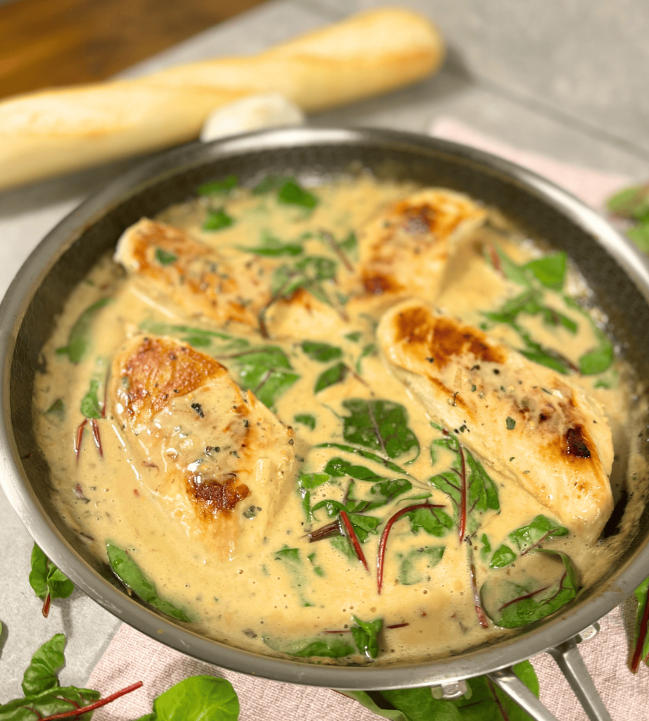Das Bild zeigt eine toskanische Hähnchenpfanne mit Mangold in einer cremig goldgelben Soße. Im Hintergrund ist eine Baguettestange zu sehen.