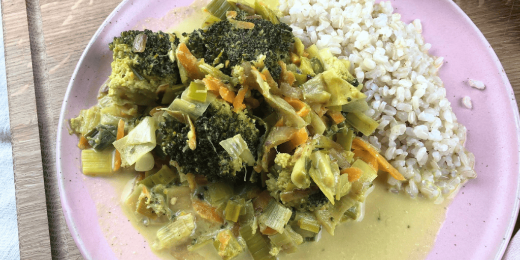 Lecker angerichtetes Curry-Gemüse auf einem rosa Teller – im Hintergrund warten Porree und Möhre auf ihren Einsatz. 