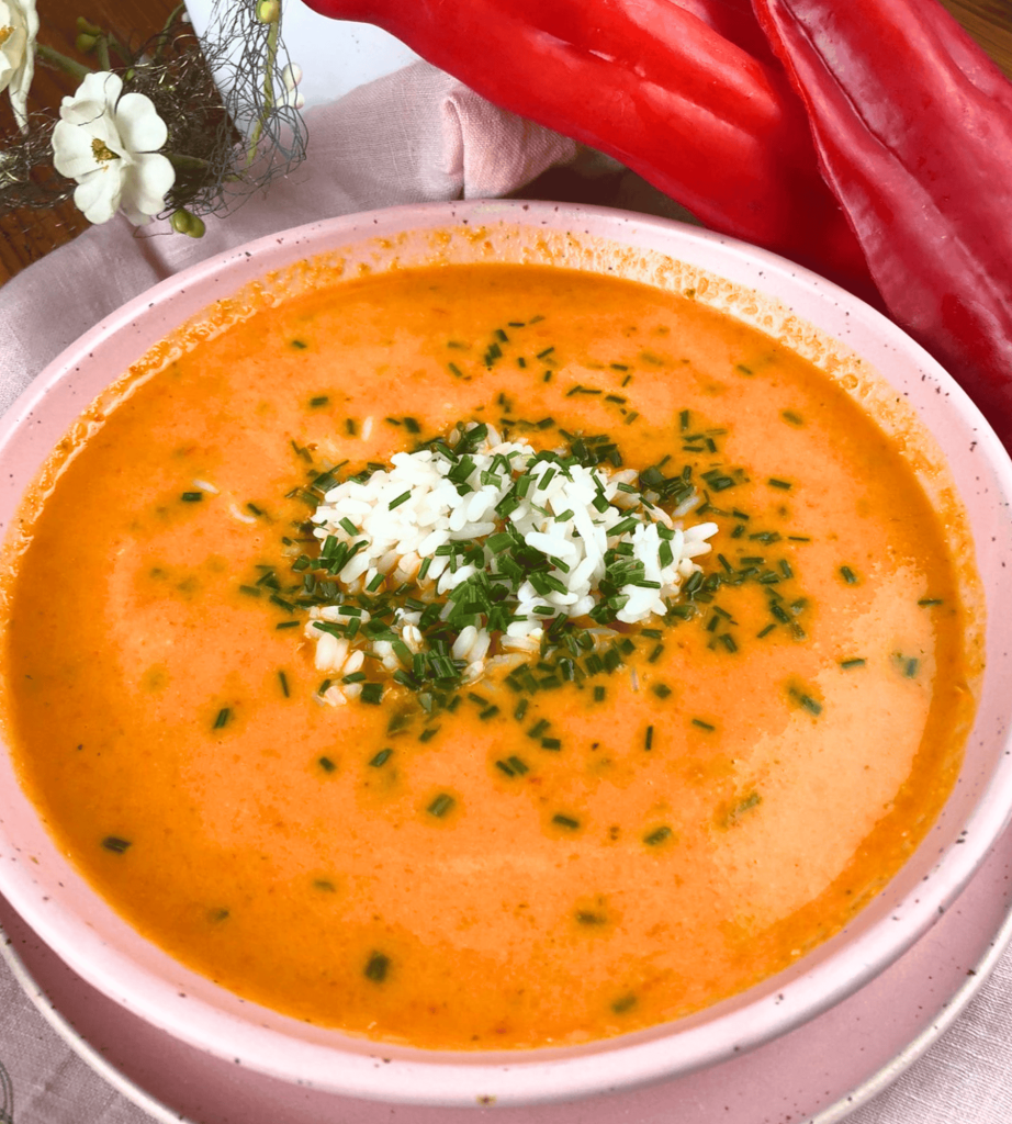 Eine cremige Paprika-Suppe, serviert in einer zarten rosa Schüssel, verfeinert mit frischen Kräutern und einer Portion Reis 