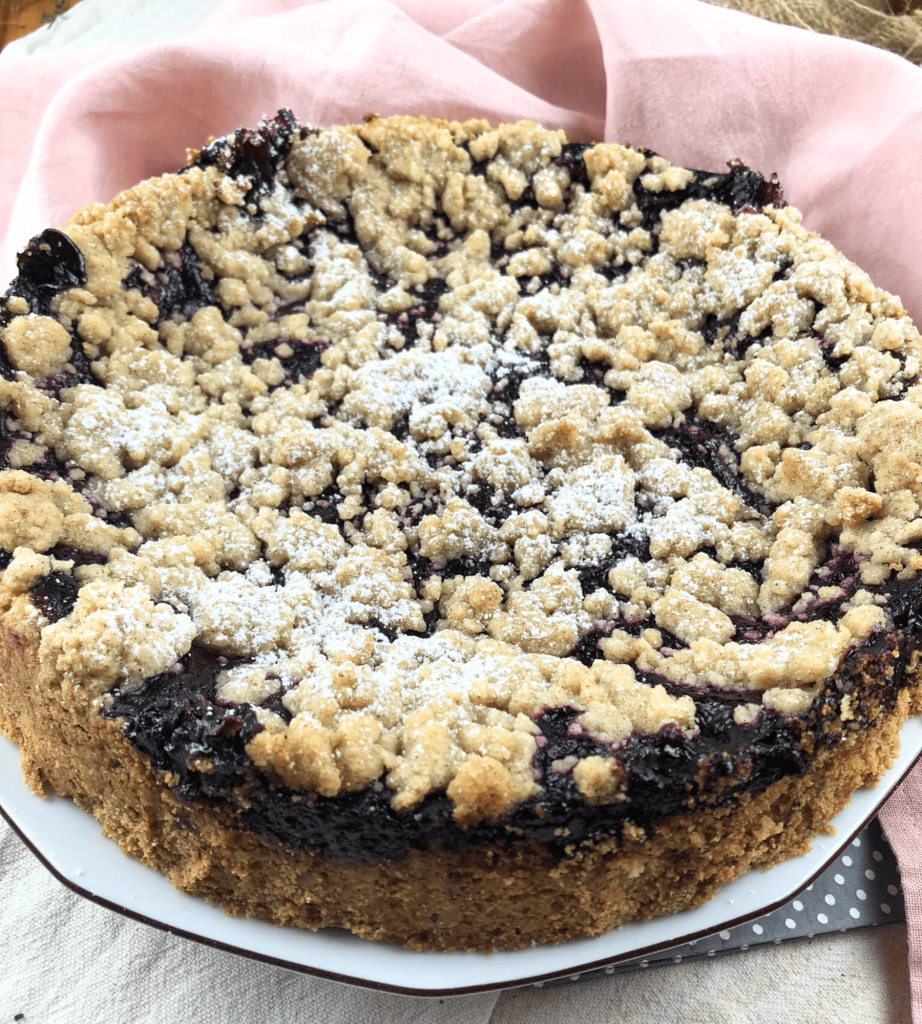 Das Bild zeigt einen frisch gebackenen Heidelbeerkuchen auf einem Teller. Der Kuchen ist mit einer feinen Schicht Puderzucker bestreut und mit knusprigen, goldbraunen Streuseln verziert. Die saftige Füllung aus Heidelbeeren schimmert leicht durch die Streusel hindurch.