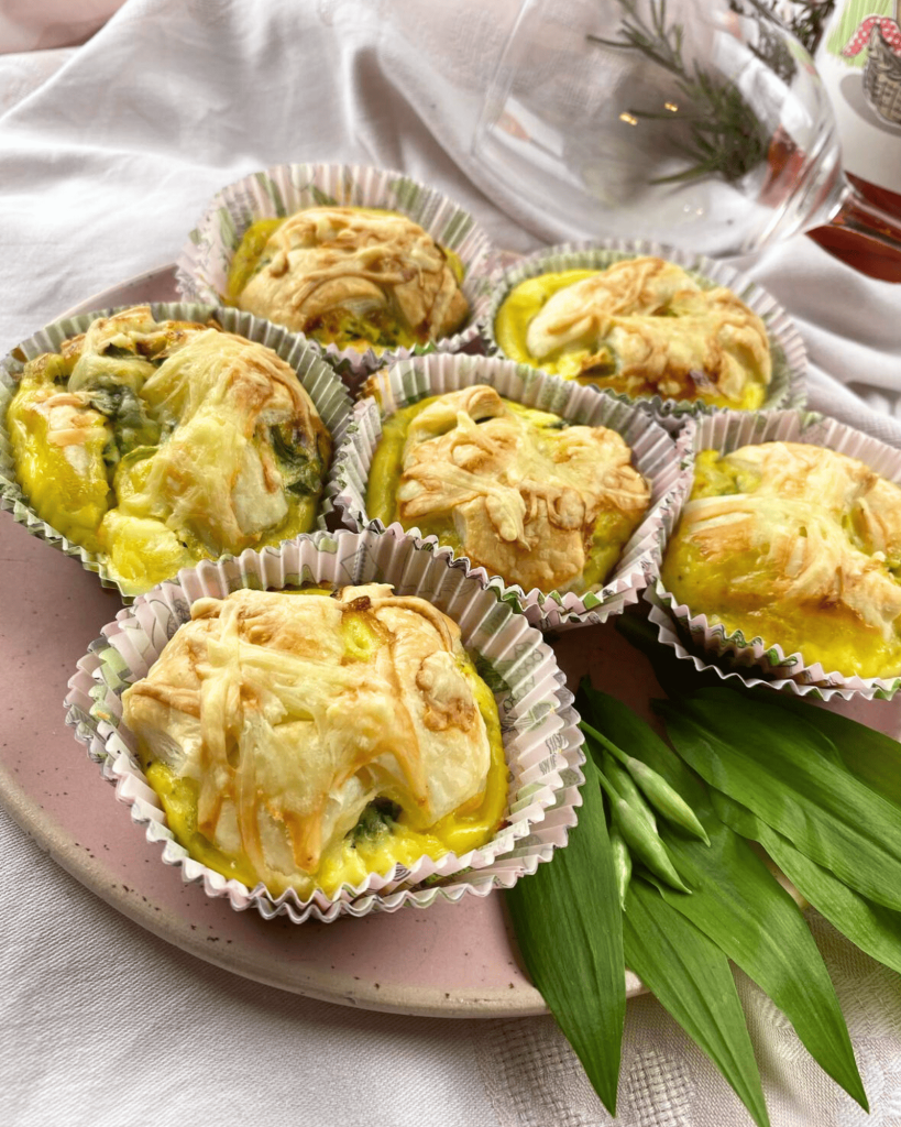 Sechs Knusprig-herzhafte Käsemuffins, serviert auf einem rosafarbenen Teller, mit goldbraunem Käse überbacken. Dekoriert mit Grünen Tulpenblättern.